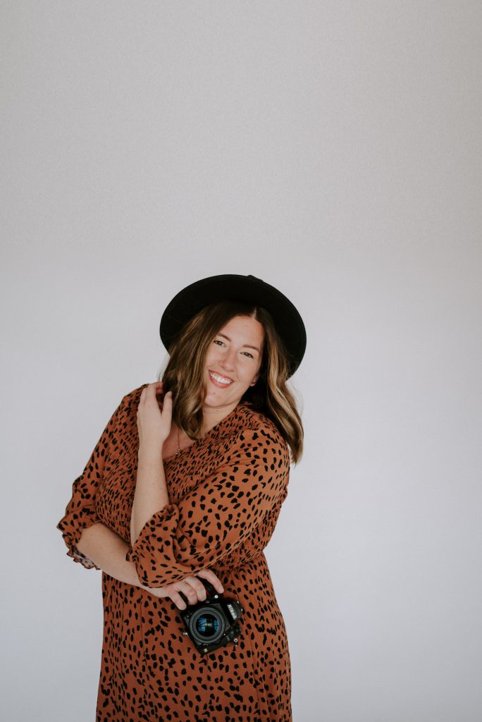 Amanda in a burnt orange dress and black hat posing with her arms crossed holding a camera and with one hand in her hair.
